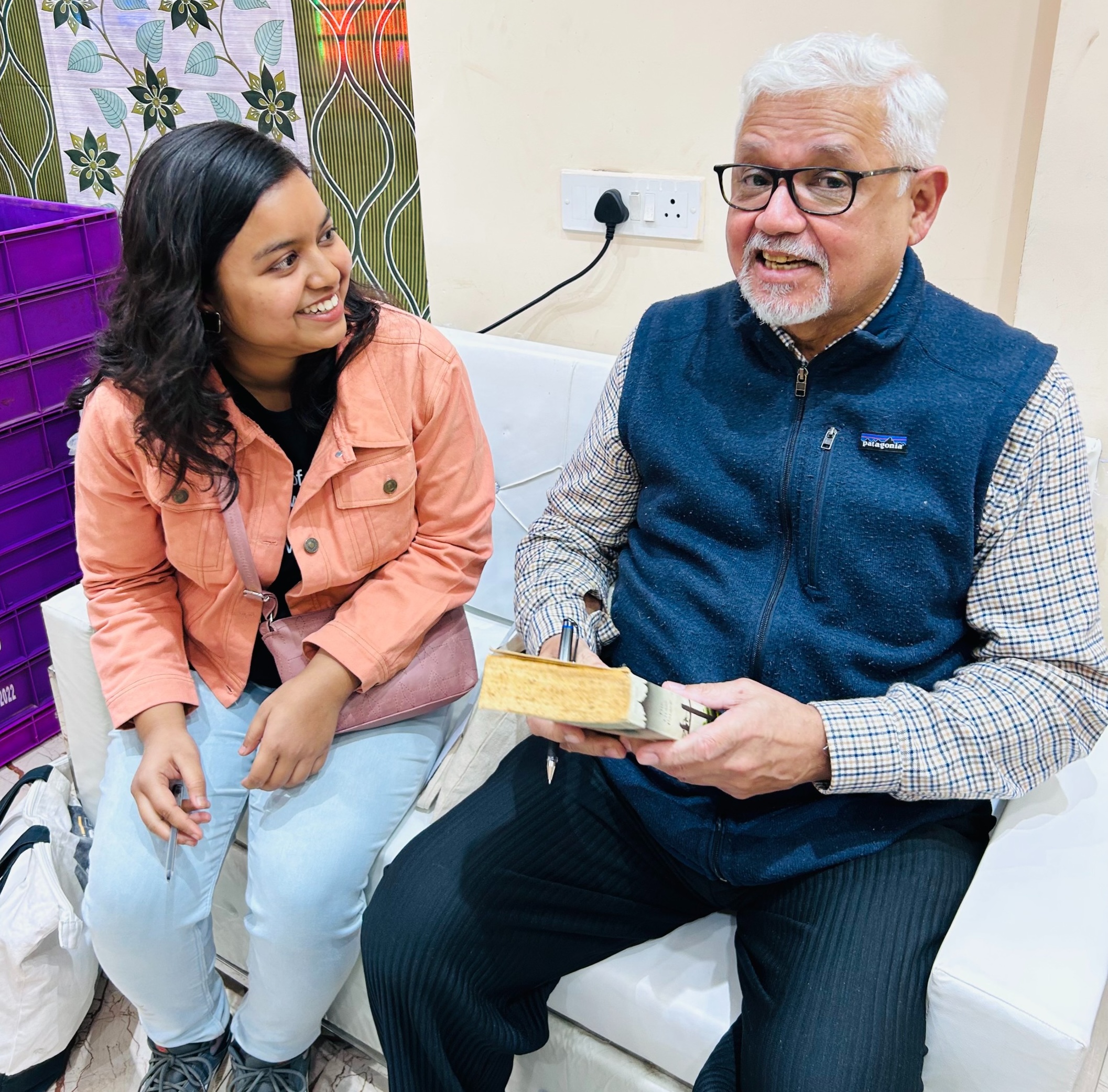 Author Amitav Ghosh sits on a white couch, holding a book and a pen while speaking. A smiling woman wearing a peach-colored jacket and light blue pants sits beside him, looking at him attentively.
