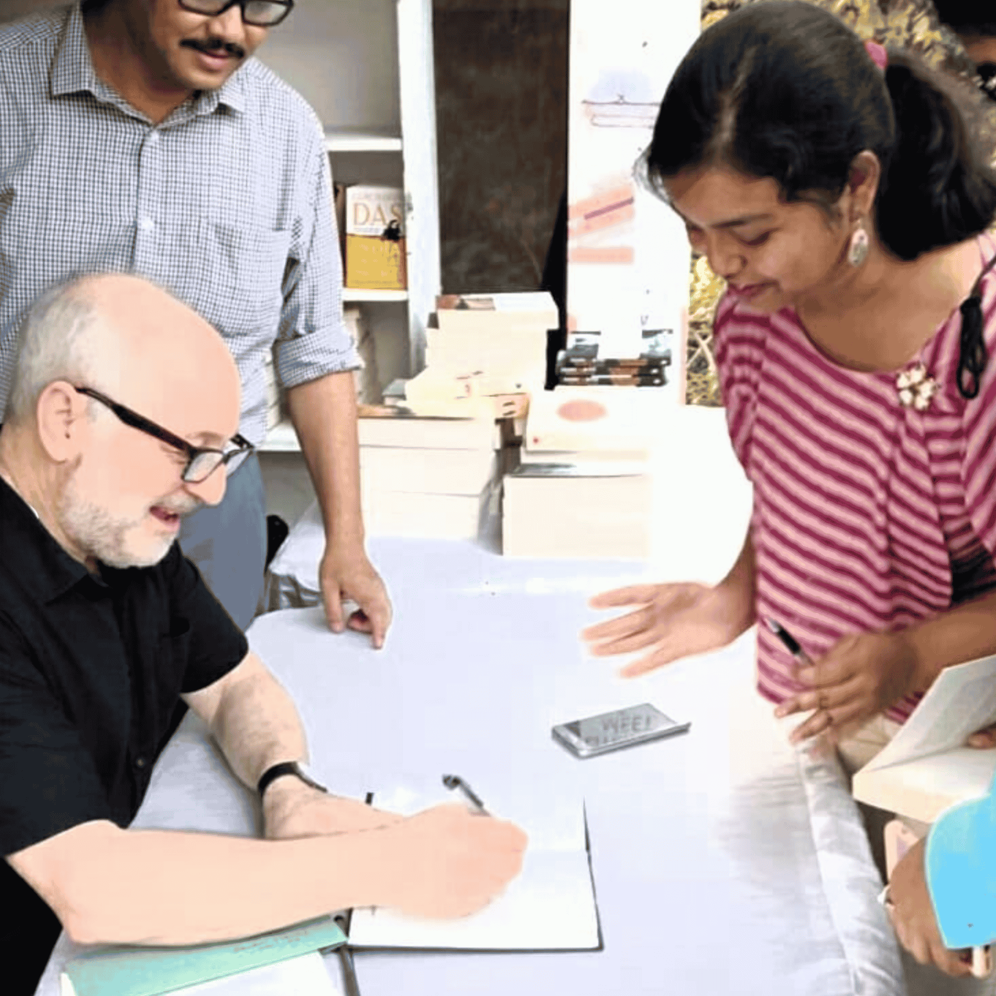 Author Andre Aciman is seated at a table, signing a book with a pen. A smiling woman in a pink and white striped top stands across from him, watching him sign. Another man stands behind the table, looking on, with stacks of books visible in the background.