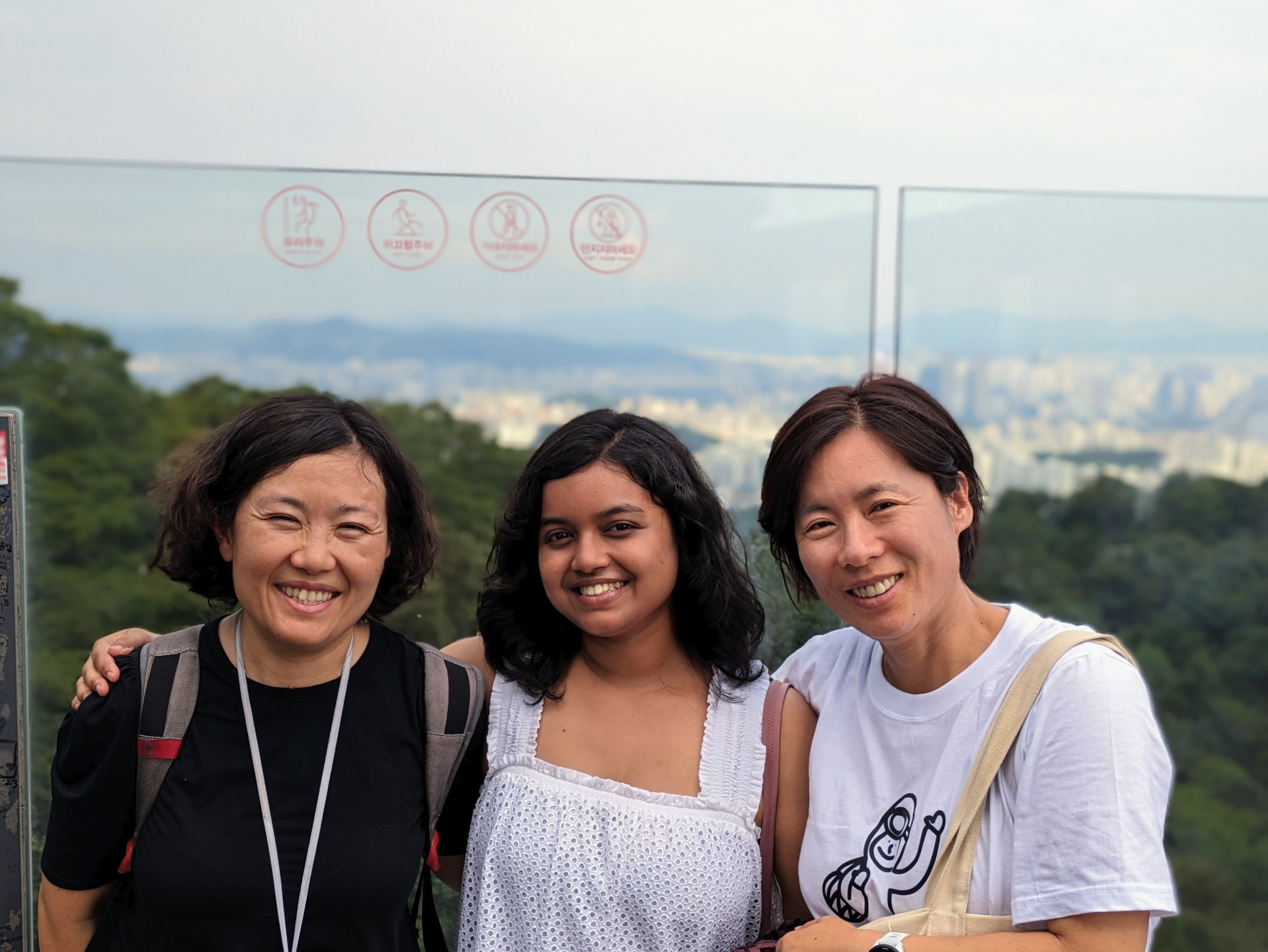 Three smiling women stand close together outdoors at an elevated viewpoint. They are positioned in front of a clear glass safety barrier that overlooks a distant city skyline nestled among rolling green hills under an overcast sky.