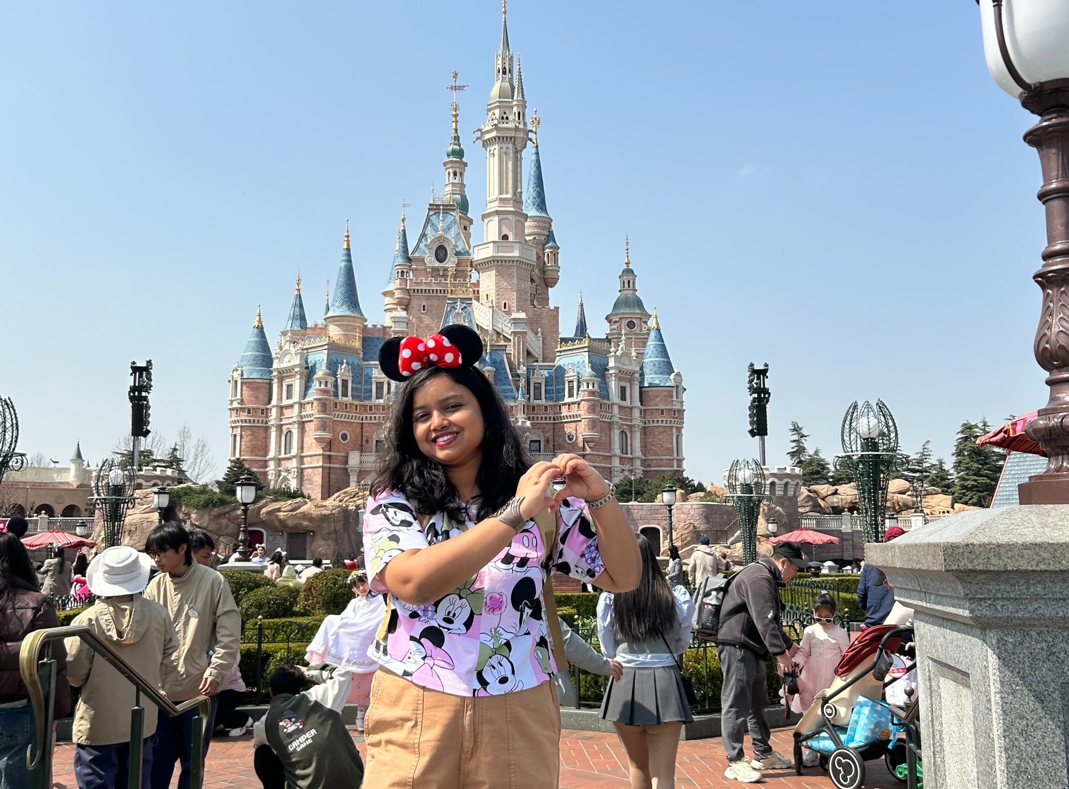 A smiling woman wearing Minnie Mouse ears and a patterned Disney shirt stands in front of a large fairy tale castle, making a heart shape with her hands. Crowds of people and park decorations are visible in the background under a clear sky.