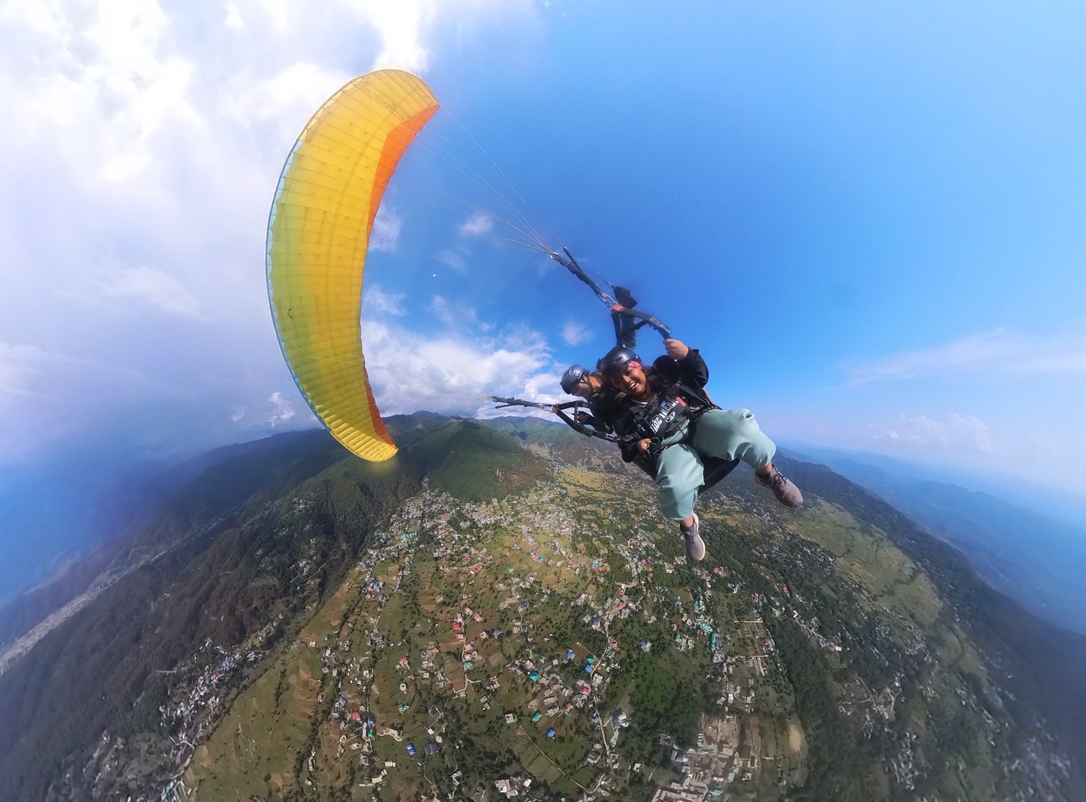 A wide-angle, birds-eye view of a tandem paragliding flight high above a valley. A smiling passenger and their pilot are suspended beneath a vibrant yellow and orange canopy, with a vast landscape of green hills and a small town visible far below under a blue sky with scattered clouds.