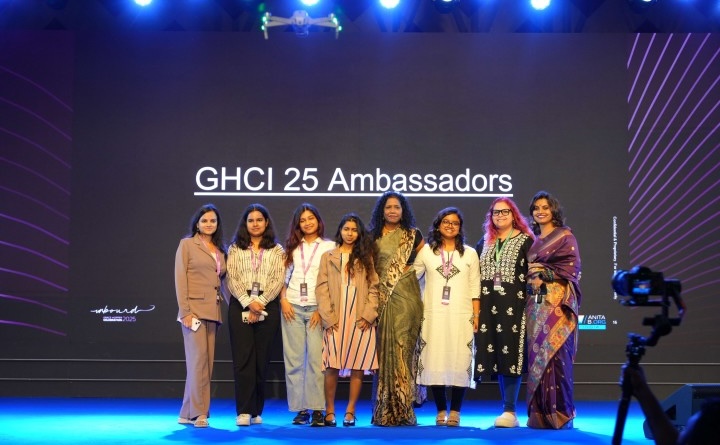 A group of eight women stand smiling together on a stage under bright event lighting. The large digital screen behind them reads 'GHCI 25 Ambassadors' in white text, with the 'Unbound Grace Hopper Celebration 2025' logo visible in the bottom-left corner. A video camera on a tripod is partially visible in the right foreground.
