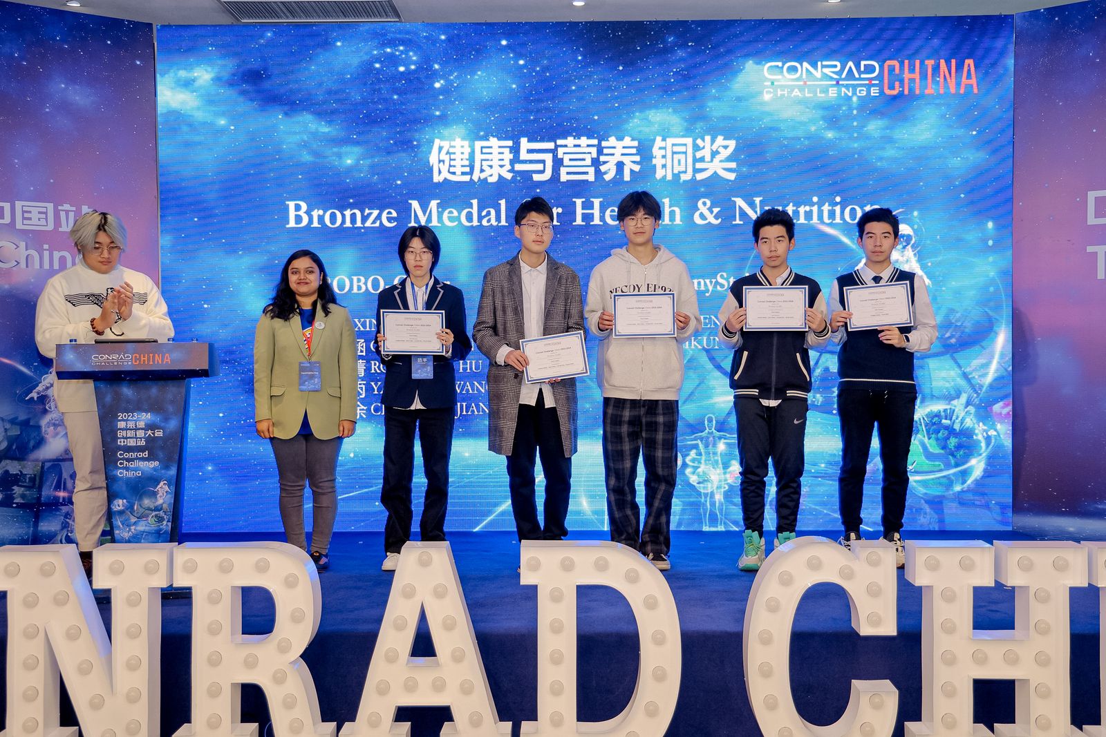 A stage photo from the ‘Conrad Challenge China’ event. Five young individuals stand in a line holding award certificates, flanked by a person standing at a podium on the far left and another woman standing next to them. The large digital screen in the background displays a starry blue galaxy with the text ‘Bronze Medal for Health & Nutrition’. Large white marquee letters spelling out ‘CONRAD CH’ are positioned in the foreground.